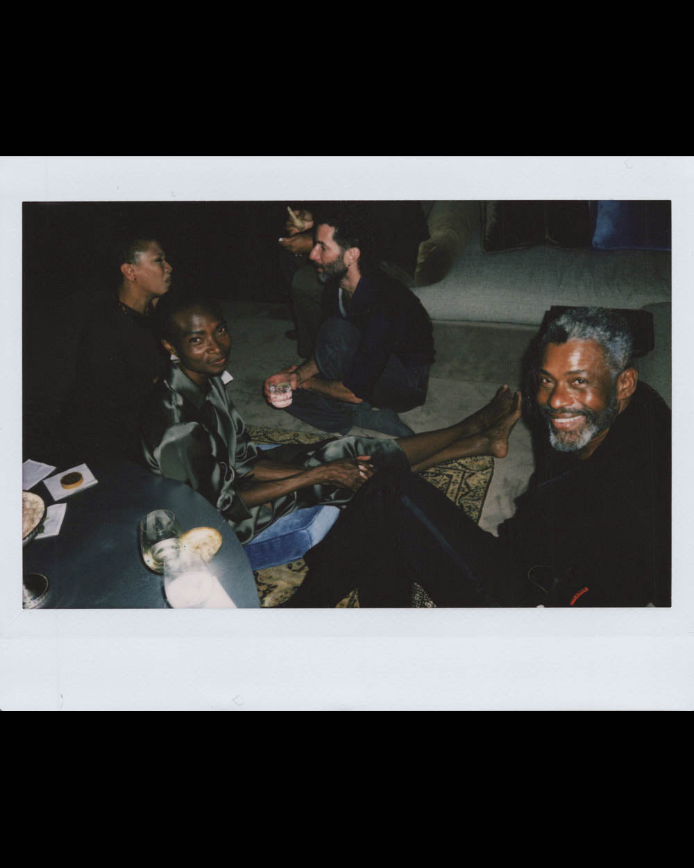 Four people sitting closely in a dimly lit room, two smiling while engaging in relaxed conversation.