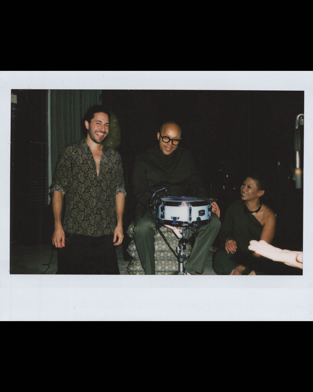 Three people smiling and interacting around a drum in a dimly lit indoor setting