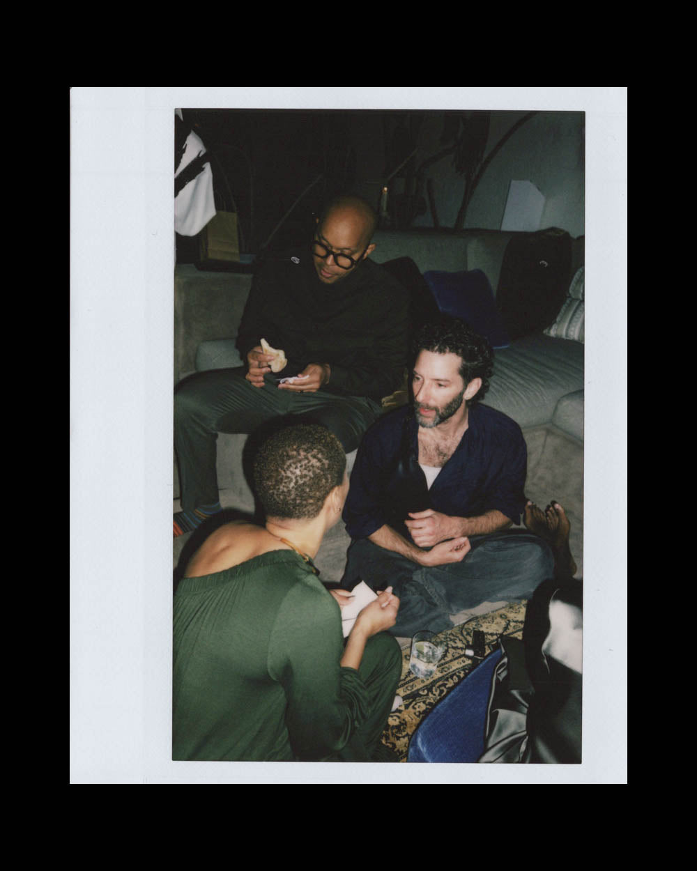 Three people engaged in conversation in a cozy room with a rug and a couch in the background.