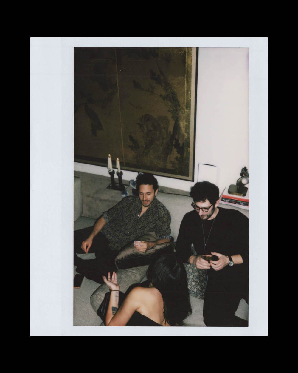 Three people seated indoors on a couch and floor, engaged in relaxed conversation in a softly lit room.