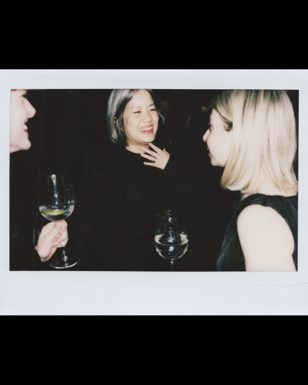 Three women holding wine glasses and smiling while chatting in a dimly lit indoor setting.