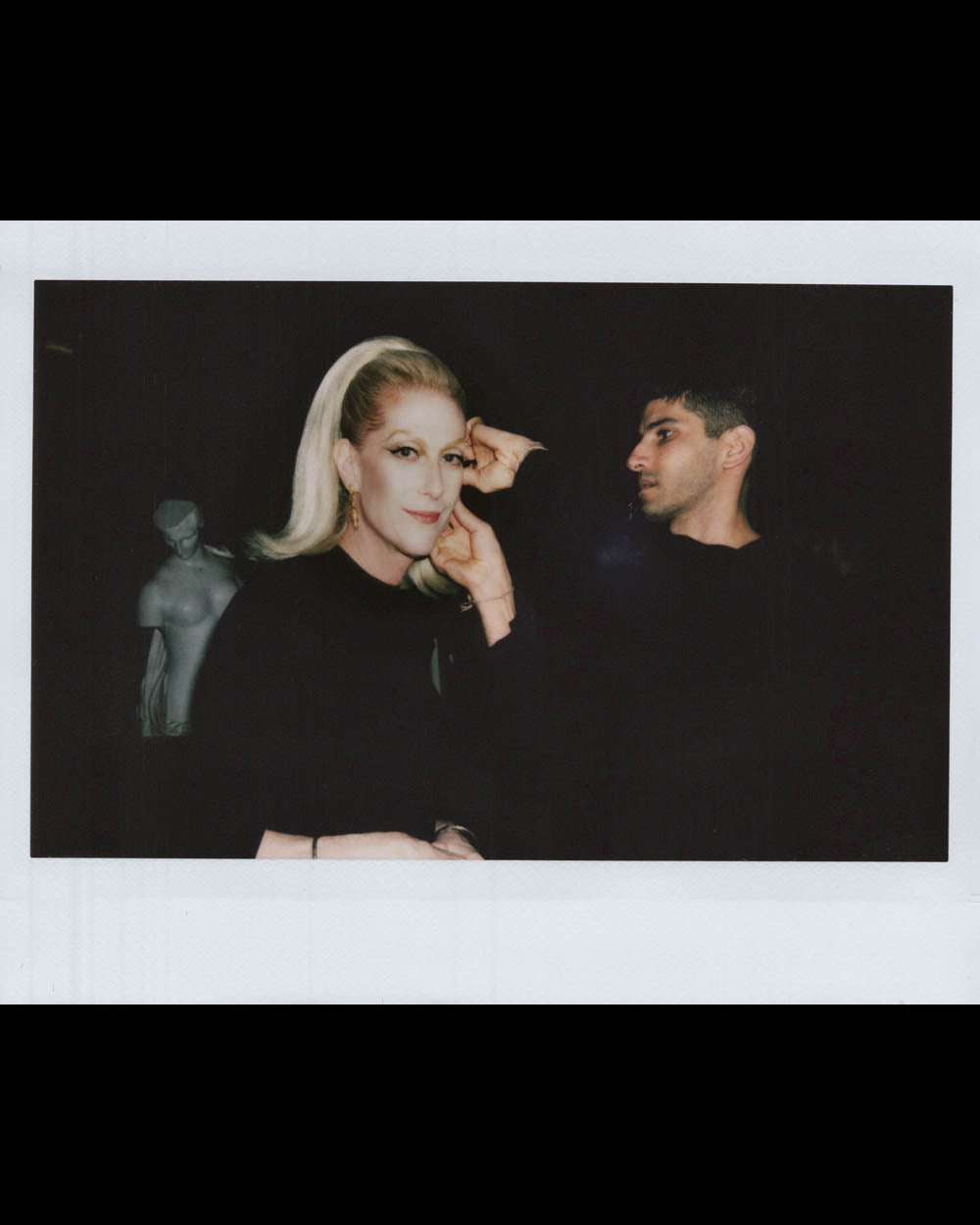 A woman with blonde hair and a man dressed in black posing indoors with a faint statue in the dark background.