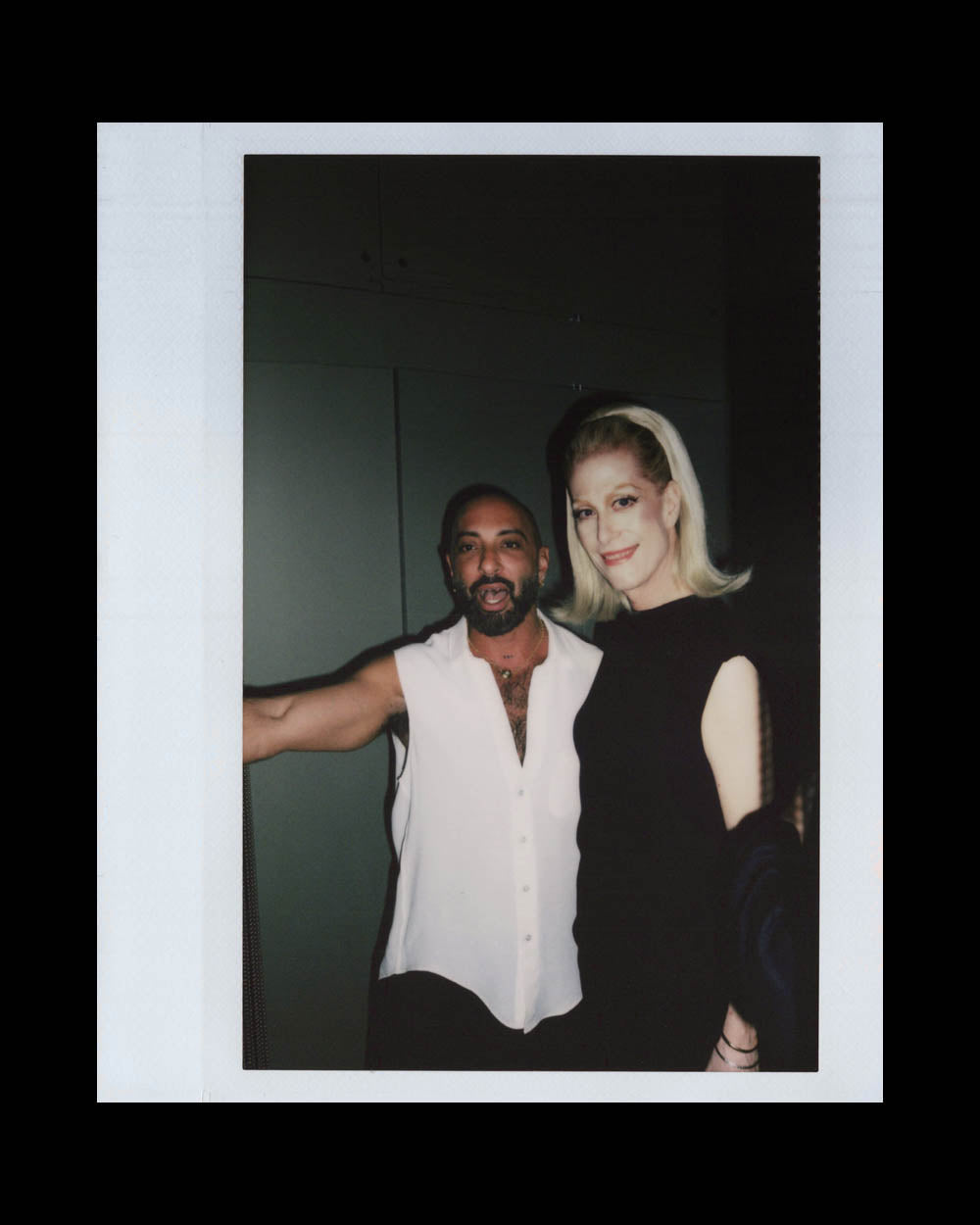 Man in sleeveless white shirt and woman in black dress posing together indoors against a dark background.