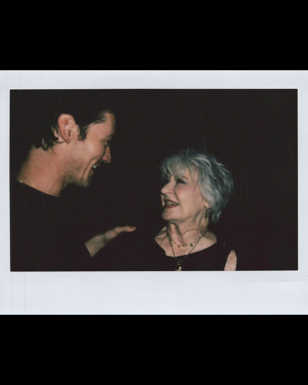 An older woman with gray hair smiling at a younger man who is gesturing with his hand in dark surroundings.