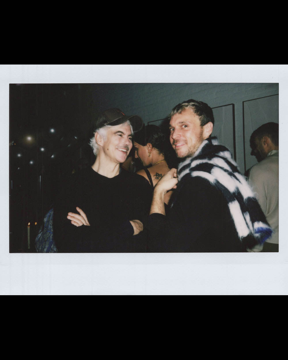 Two men facing each other and smiling in a dimly lit indoor space with people in the background.