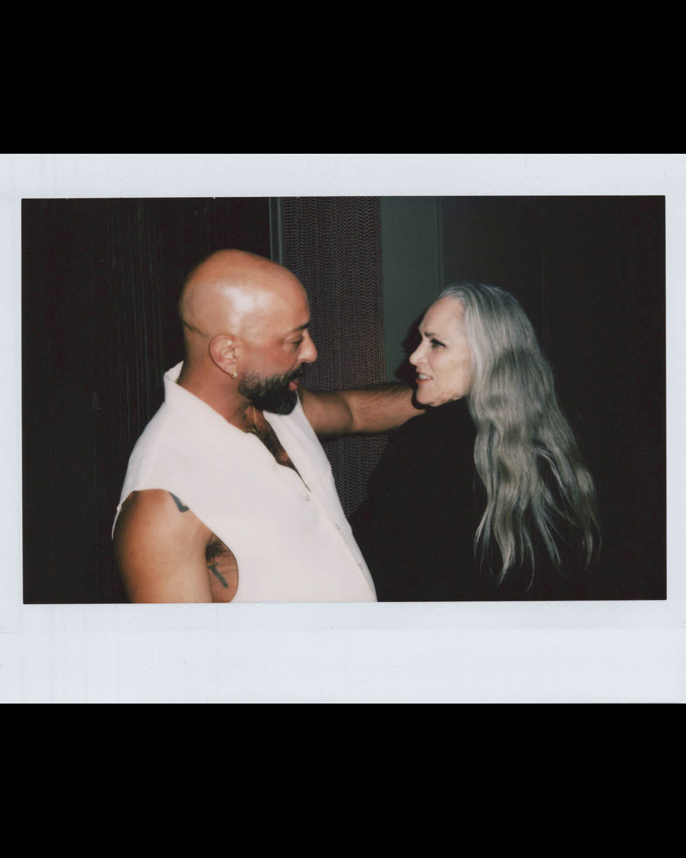 A bald man in a sleeveless white shirt embraces a woman with long gray hair against a dark background.