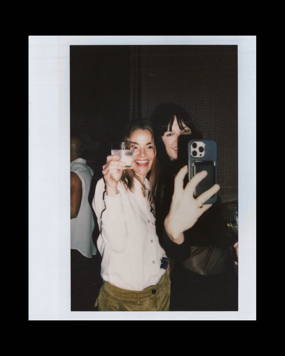 Two women smiling and taking a selfie together in a dimly lit indoor setting, one holding a drink.