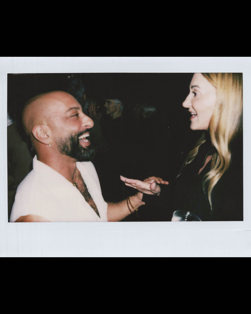 Man with beard and woman with long blonde hair laughing and talking in a dark indoor setting.