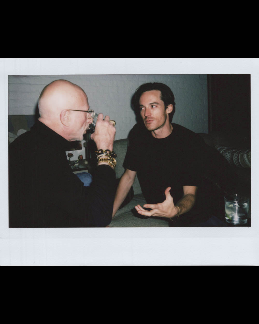 Two men sitting and talking indoors, one with glasses drinking from a glass, the other gesturing with his hand.