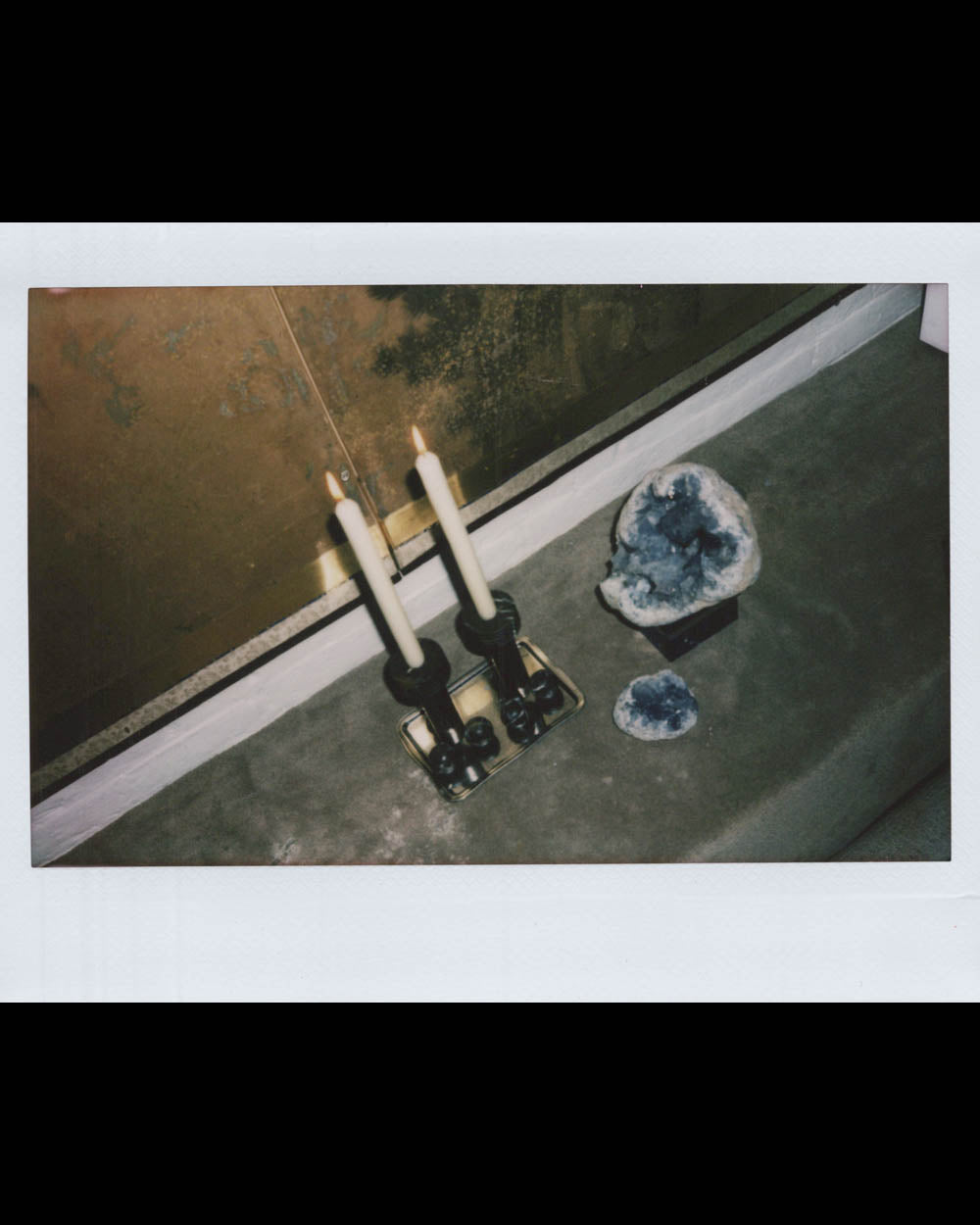 Two lit candles in black holders on a gray surface next to a large and small blue geode against a brown wall.