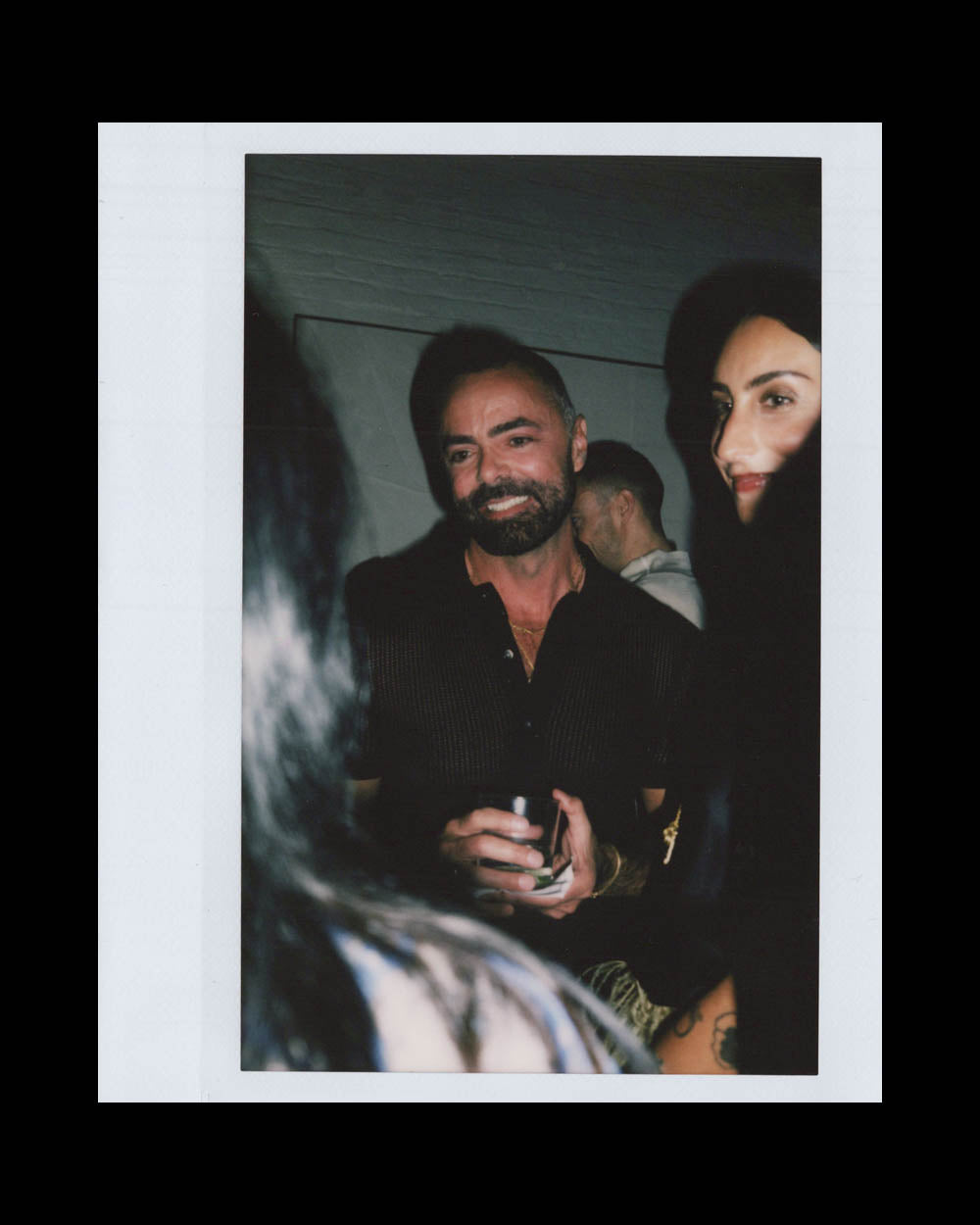 Man with a beard holding a drink and smiling among people in a dimly lit indoor setting