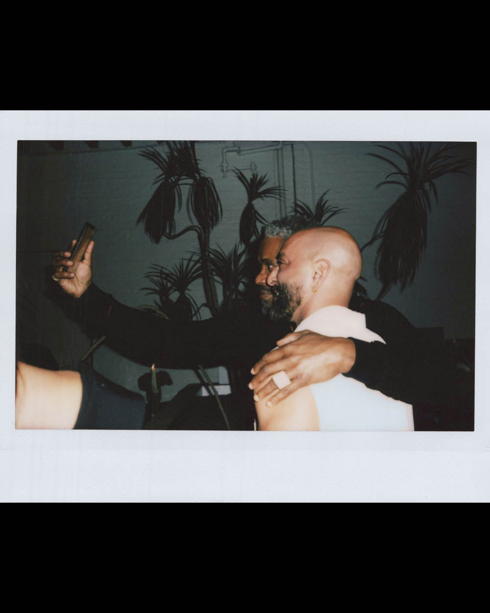 Two men closely posing for a selfie in a dimly lit room with dark plant silhouettes in the background