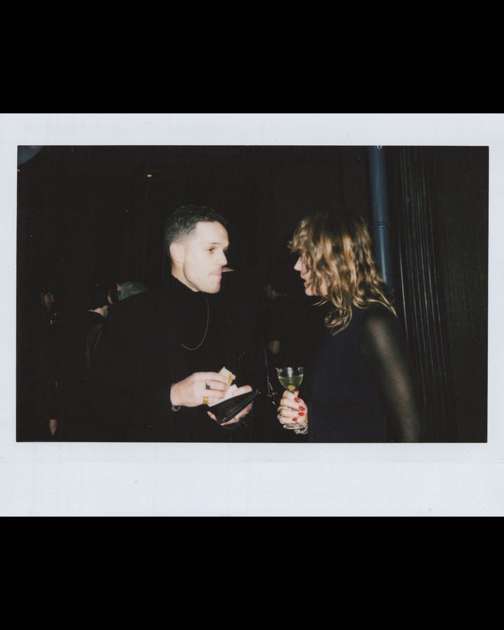A man and woman conversing indoors, the man holding a plate and the woman holding a glass with a dark background.