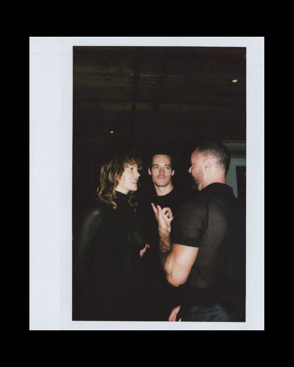 Three people engaged in conversation indoors, two men and one woman, all wearing dark clothing.