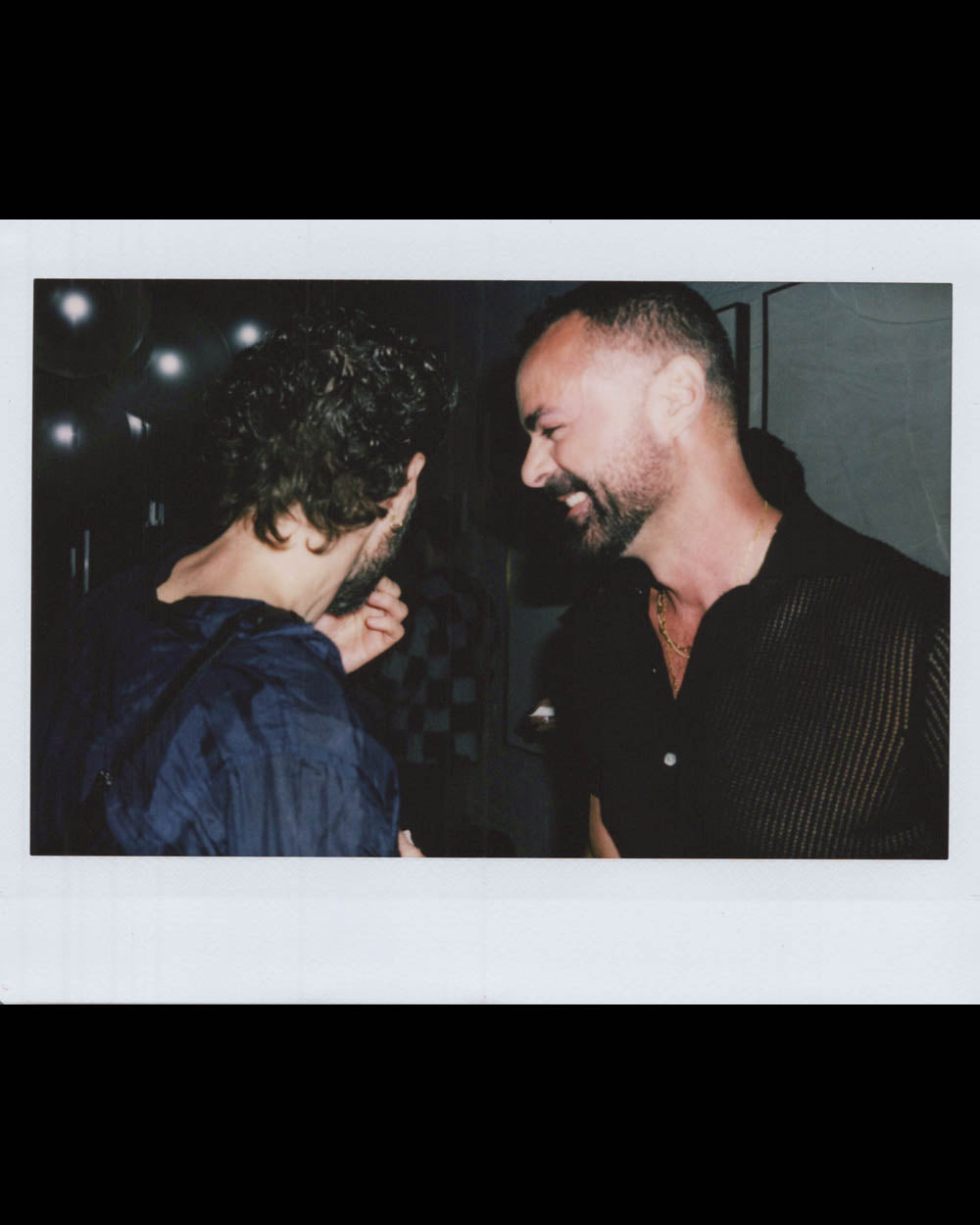 Two men sharing a laugh indoors, one facing sideways and the other looking down with a smile.
