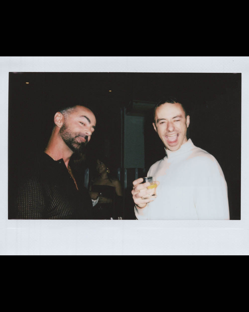 Two men in dim lighting, one with a beard looking down, the other in white laughing while holding a drink