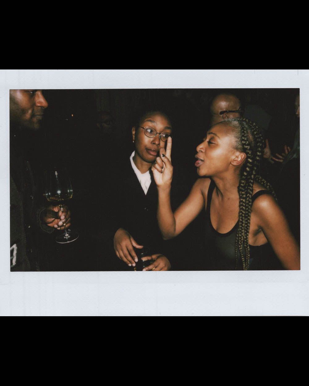 A woman with braided hair animatedly speaking to a woman wearing glasses while a man nearby holds a wine glass.