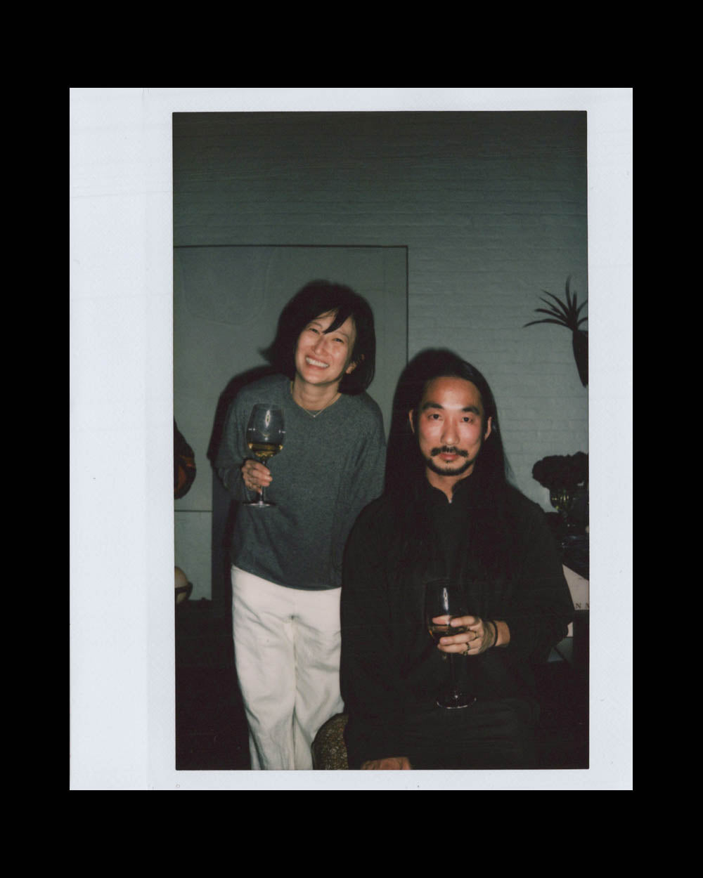 A smiling woman standing behind a seated man, both holding wine glasses in a dimly lit room.