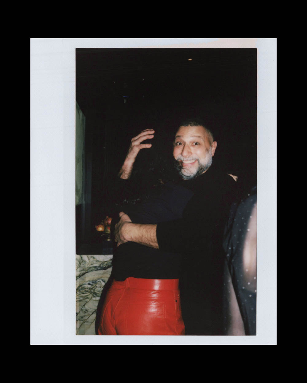 Man with gray beard smiling and hugging person wearing red leather pants in a dimly lit room