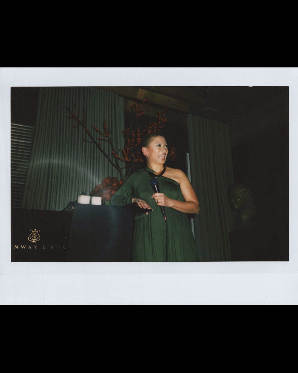 A woman in a dark green dress holding a microphone, standing indoors next to a table with candles and decorative branches.
