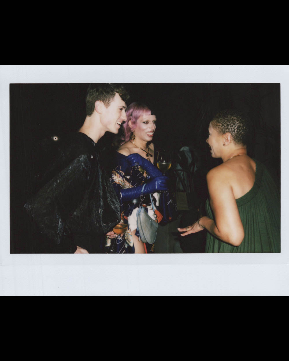 Three people in dark setting talking, one with short purple hair and blue gloves holding a drink, two others smiling and engaged