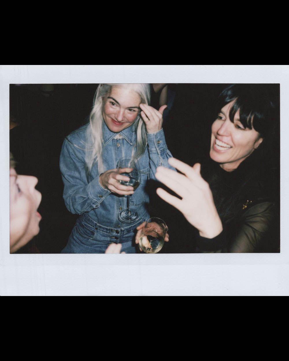 Three women smiling and holding drinks while engaging in lively conversation indoors at night.