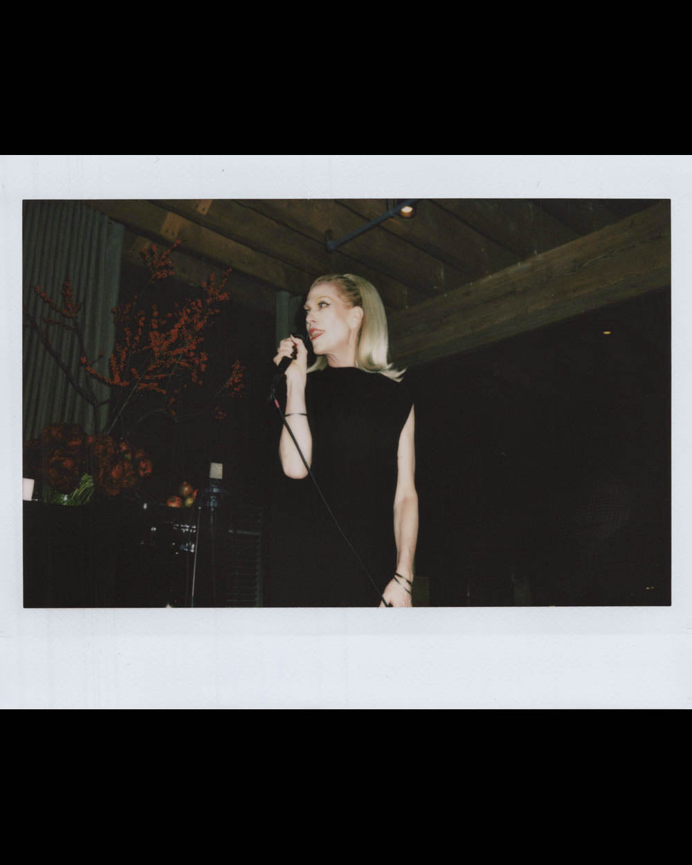 A woman with blonde hair wearing a black sleeveless dress holding a microphone indoors under wooden beams.