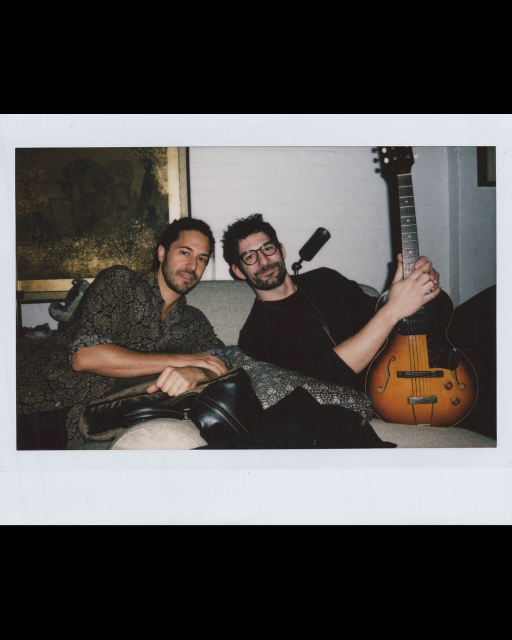 Two men sitting on a couch, one holding an electric guitar, both relaxed in a cozy indoor setting.