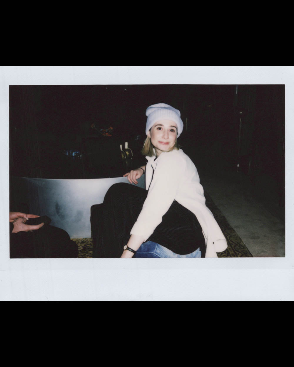 Young woman wearing a white beanie and jacket sitting on a step in a dimly lit indoor space.