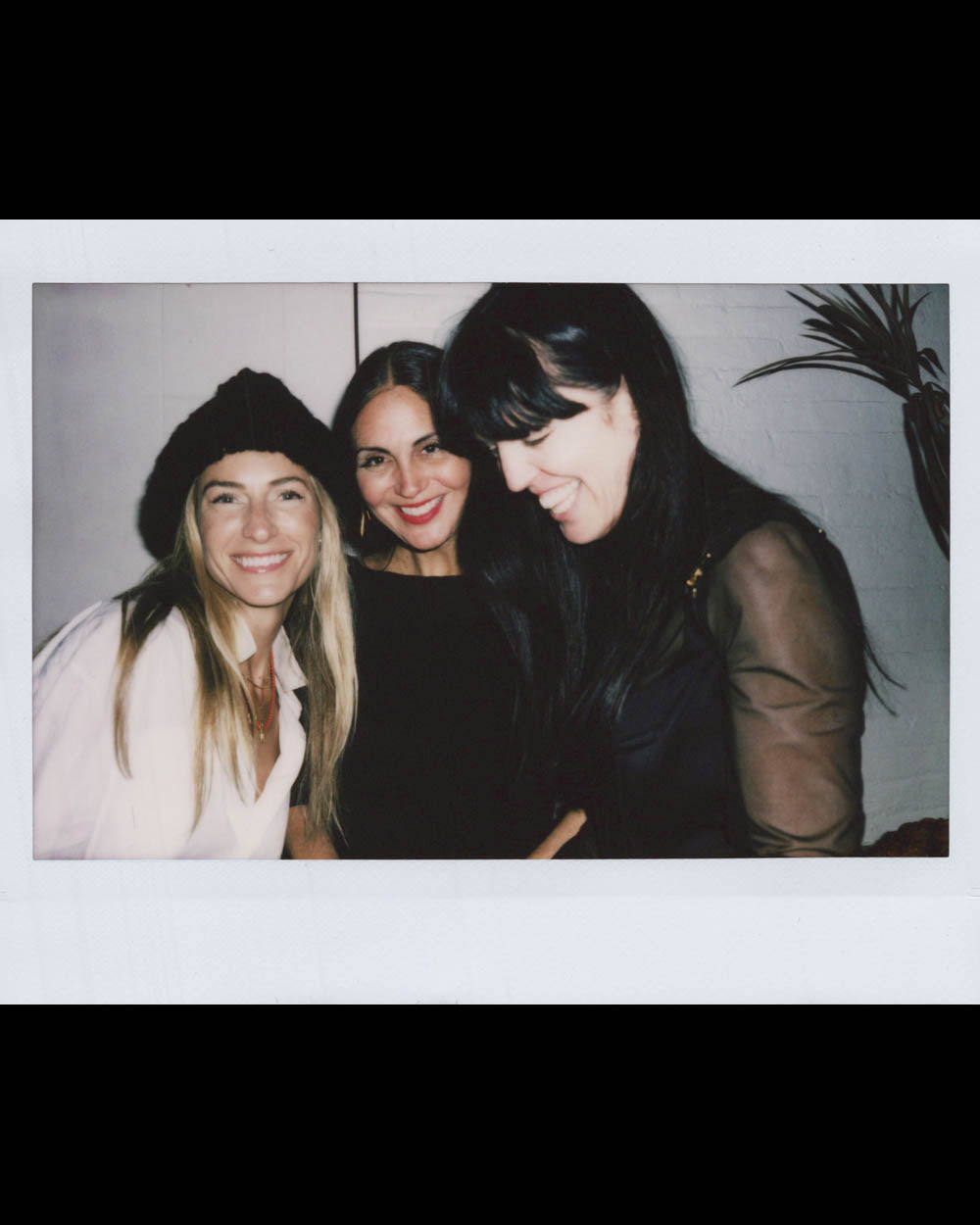 Three women smiling and posing closely in casual indoor setting with a plant visible in the background.