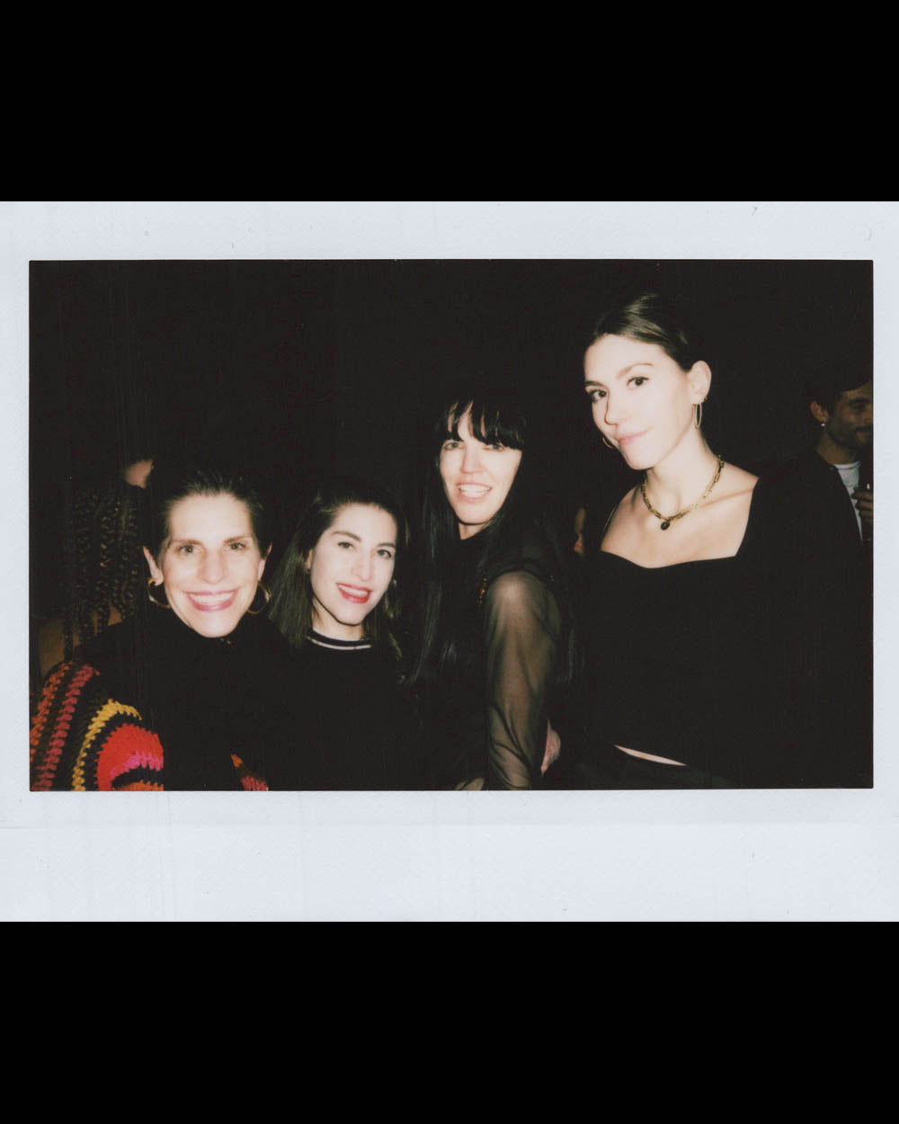 Four women smiling and posing closely together against a dark background in a casual setting
