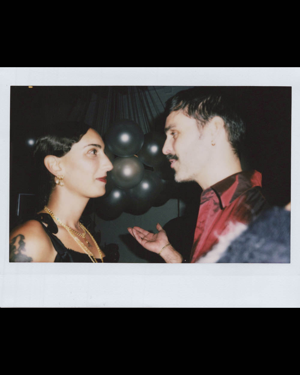 A woman and man face each other closely, in conversation, with dark balloons visible in the background.