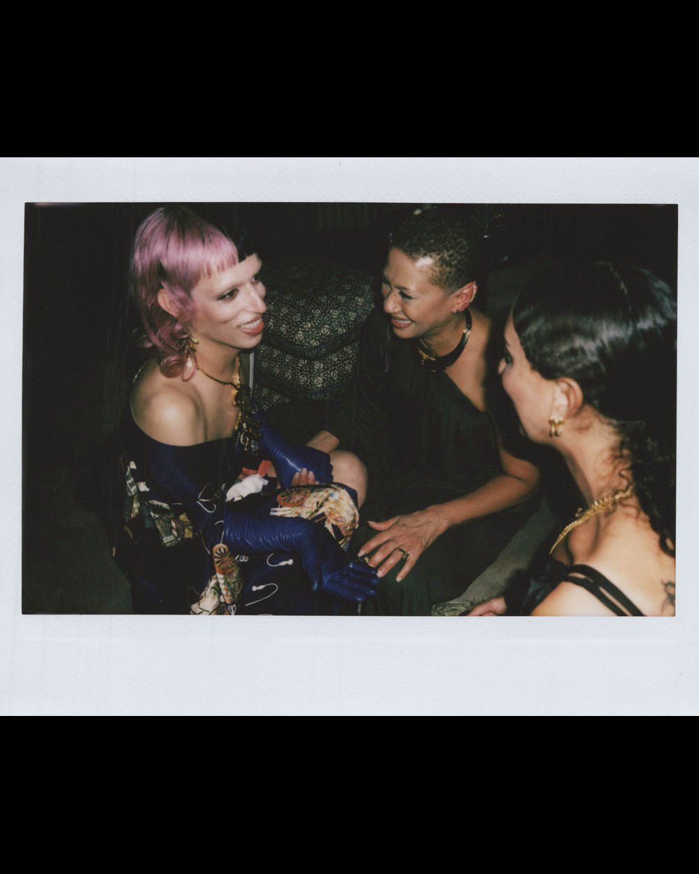 Three women seated closely indoors, smiling and engaged in conversation in a warmly lit setting
