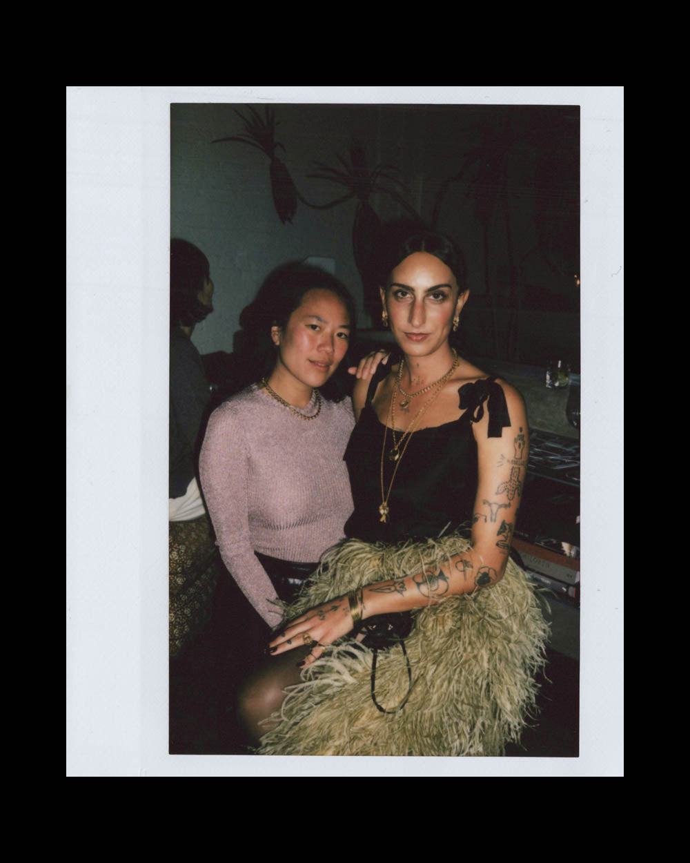 Two women posing closely indoors; one wears a purple sweater, the other a black top and textured skirt with tattoos.