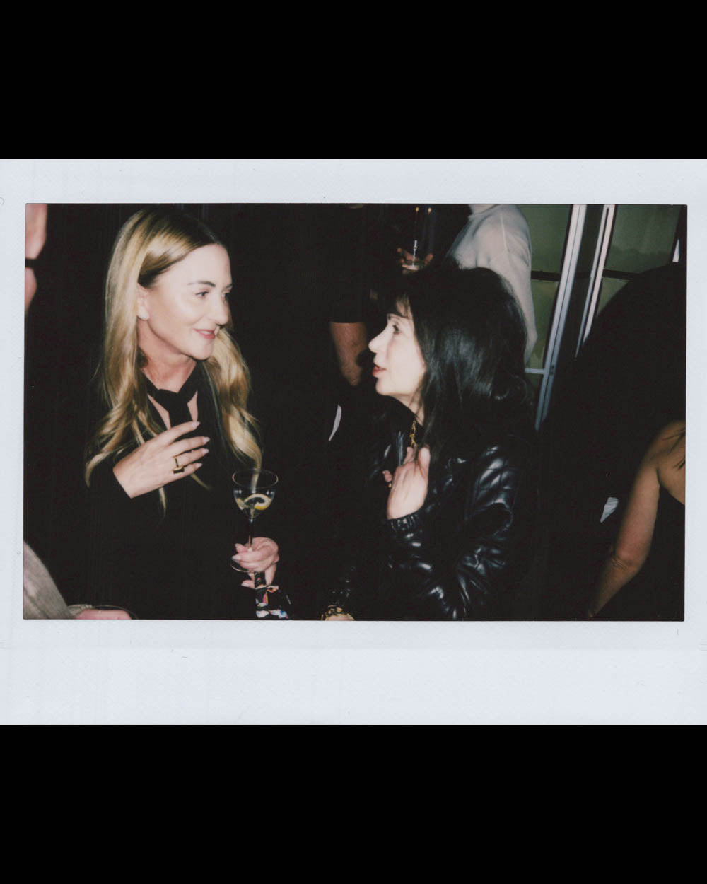 Two women engaged in conversation at a dimly lit indoor social gathering, one holding a glass.