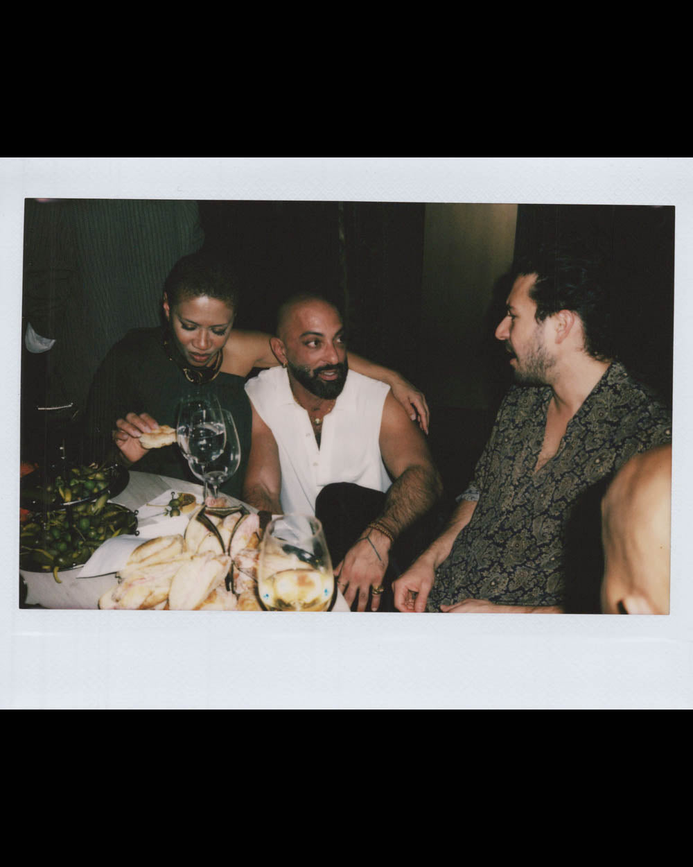 Three people seated at a table with food and drinks in a dimly lit indoor setting, engaged in conversation.