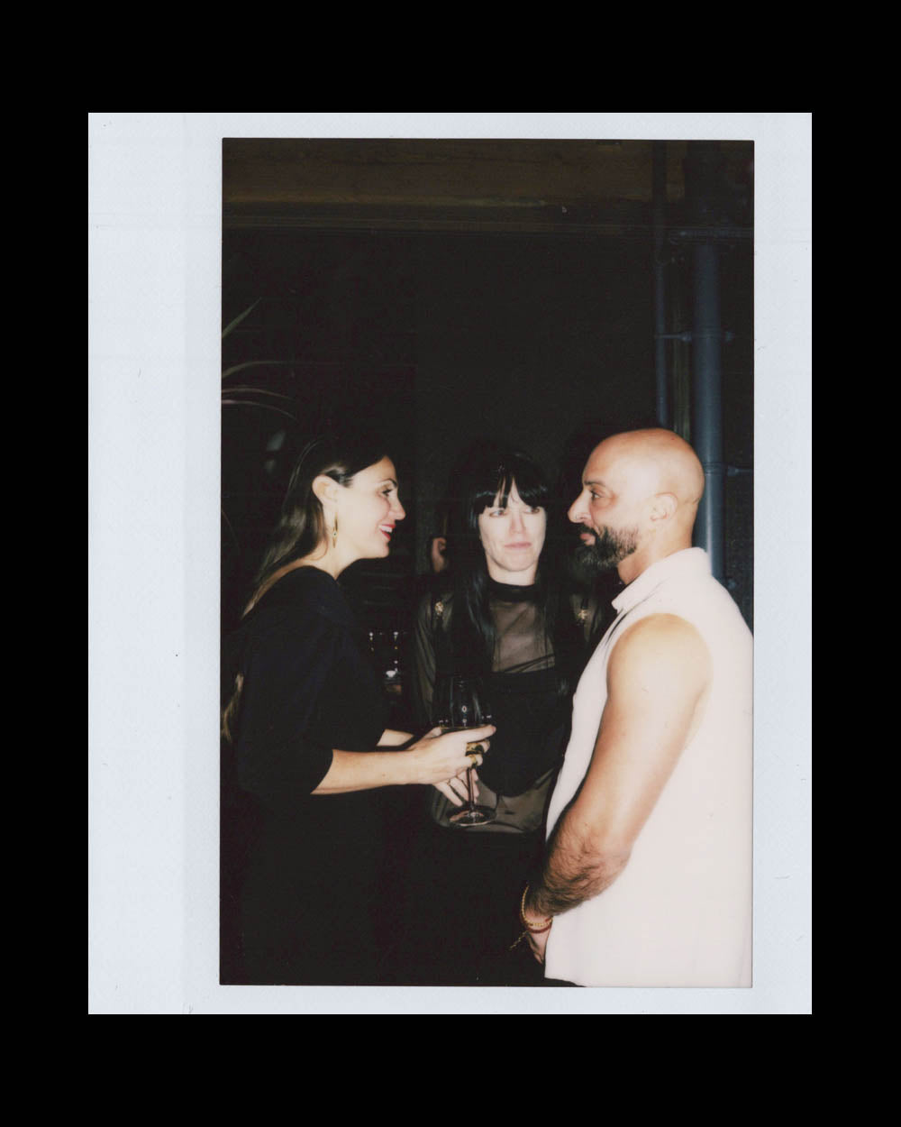 Three people standing close together in a dimly lit space, two women and a bald man in a sleeveless top.