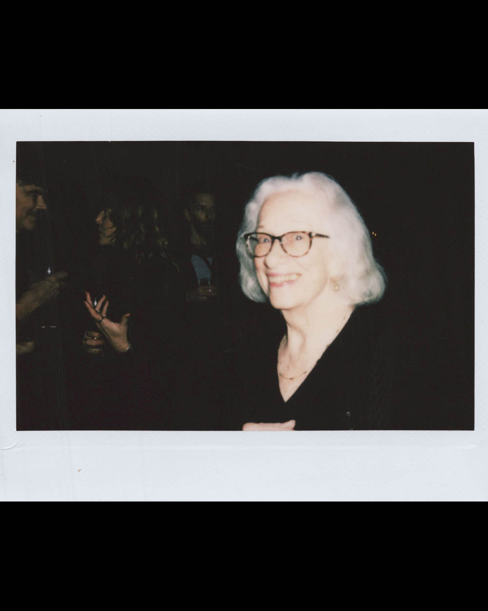 An elderly woman with white hair and glasses smiling in a dimly lit room with several people blurred in the background.