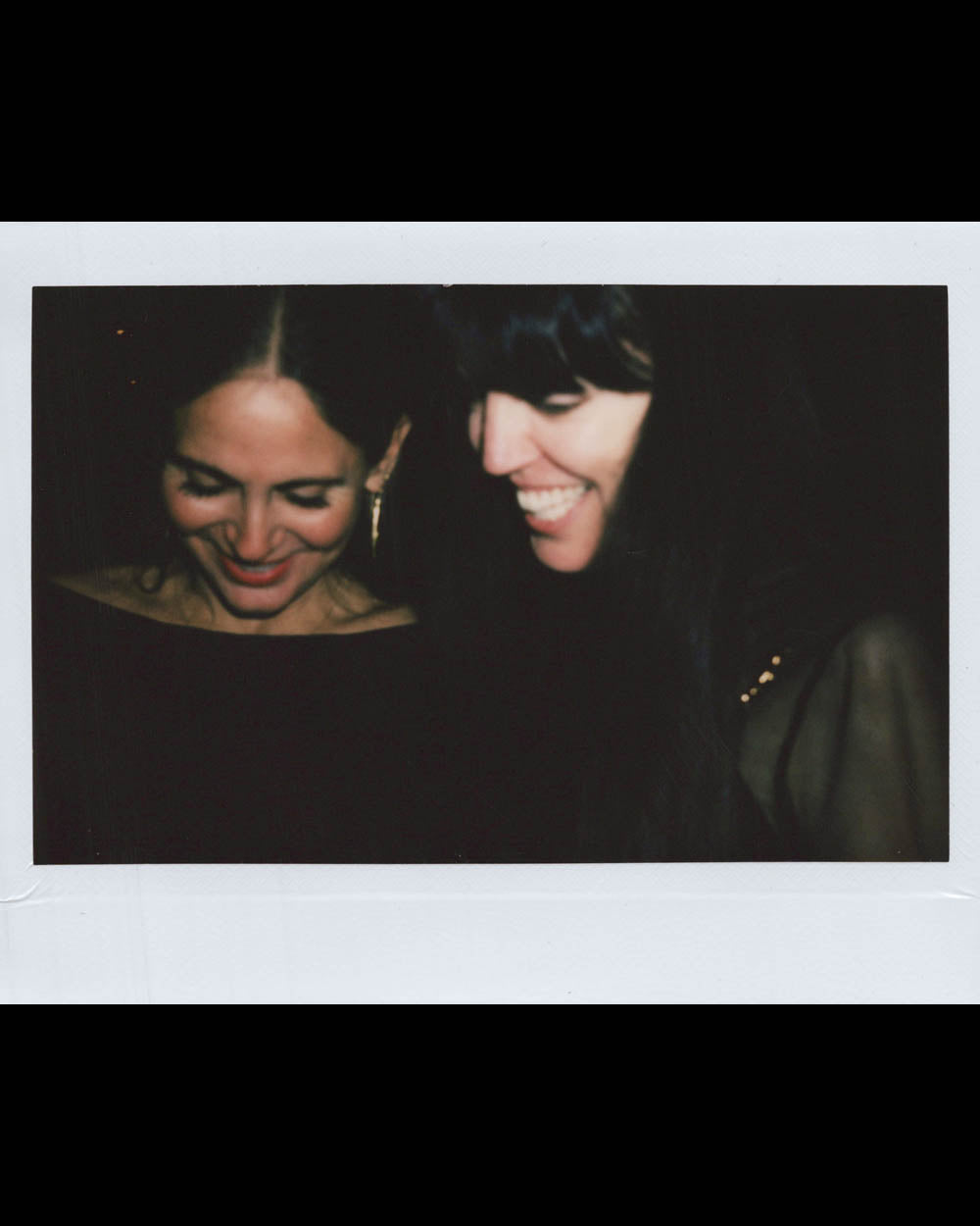 Two women leaning close and smiling while looking down in a dimly lit setting with dark clothing.