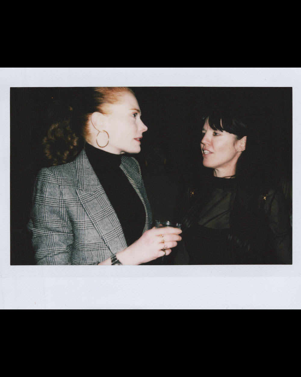 Two women engaged in conversation in a dimly lit setting, one holding a drink and wearing a plaid blazer.