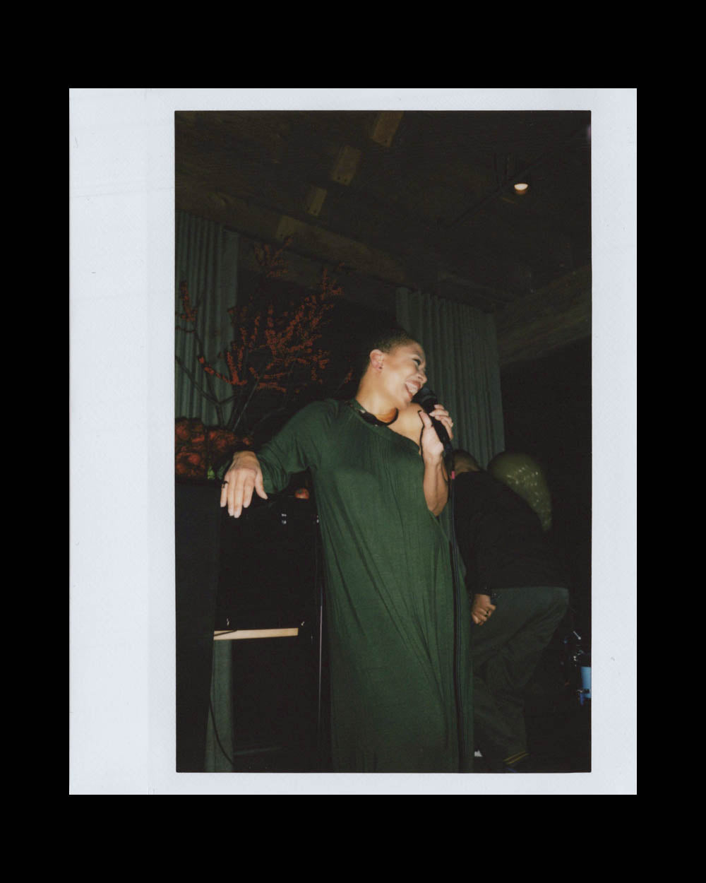 A woman in a long dark green dress holding a microphone and smiling in a dimly lit indoor space.