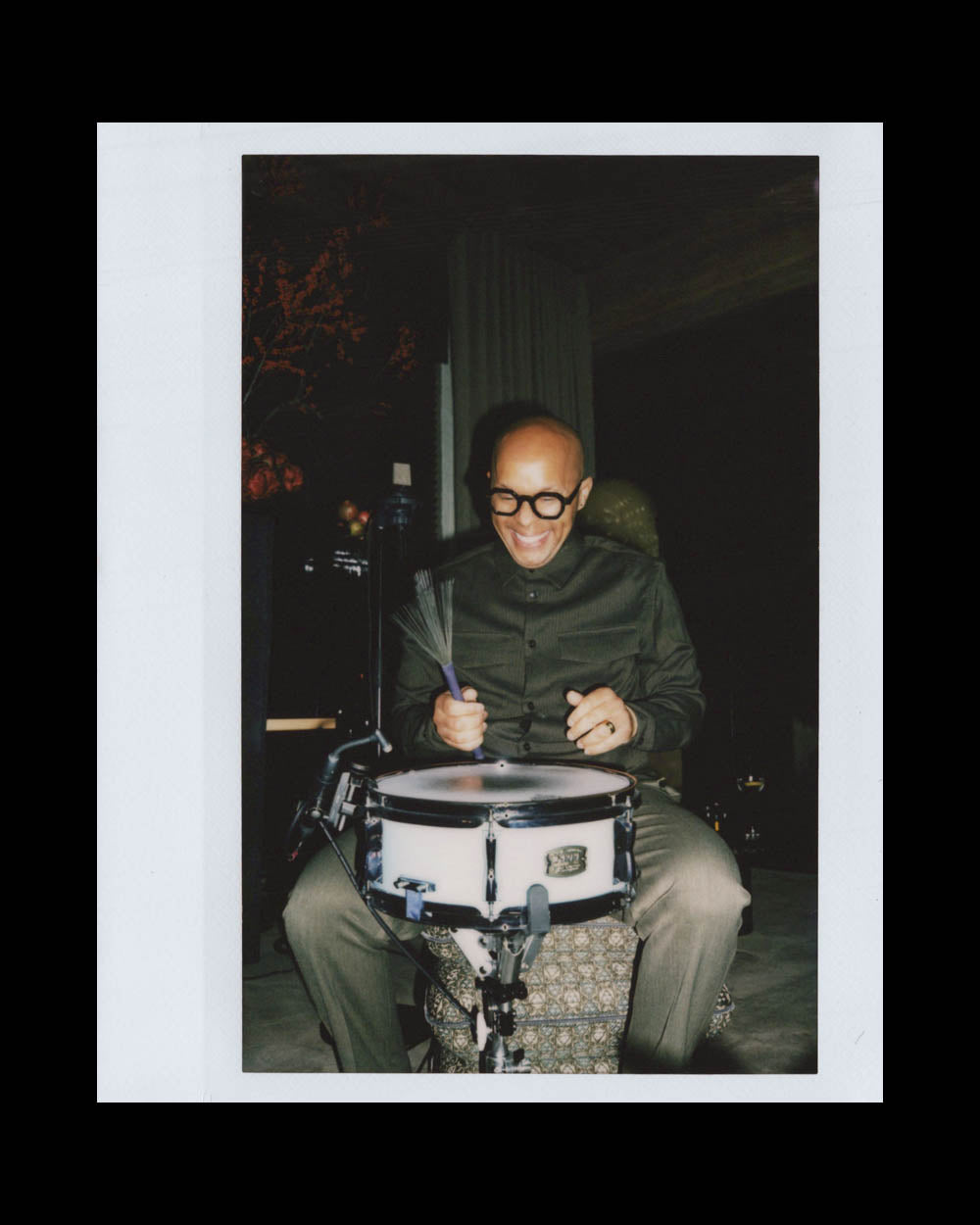 A bald man wearing round glasses and a dark shirt smiling while playing a snare drum indoors.
