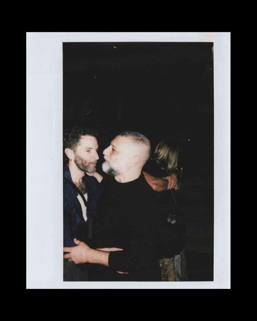 Two men face each other closely, one embracing the other against a dark background with a partially visible person behind them.