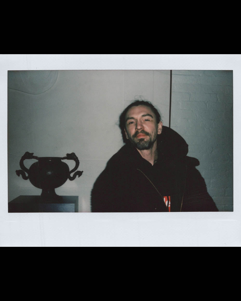 A man with a beard and tied-back hair wearing a dark jacket poses indoors next to a dark sculpture on a pedestal.