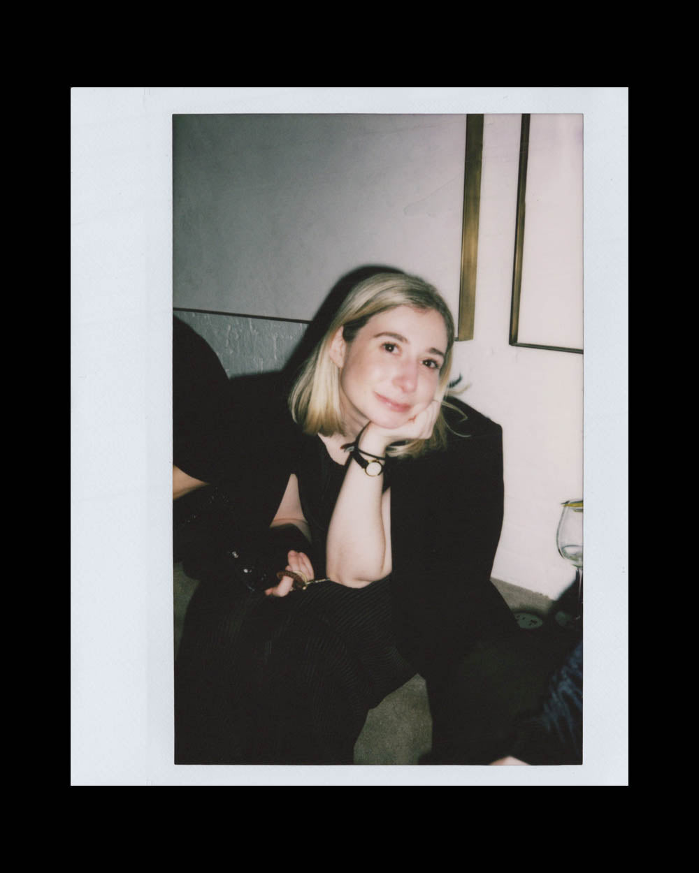 A young woman with blonde hair resting her chin on her hand, sitting indoors against a light wall.