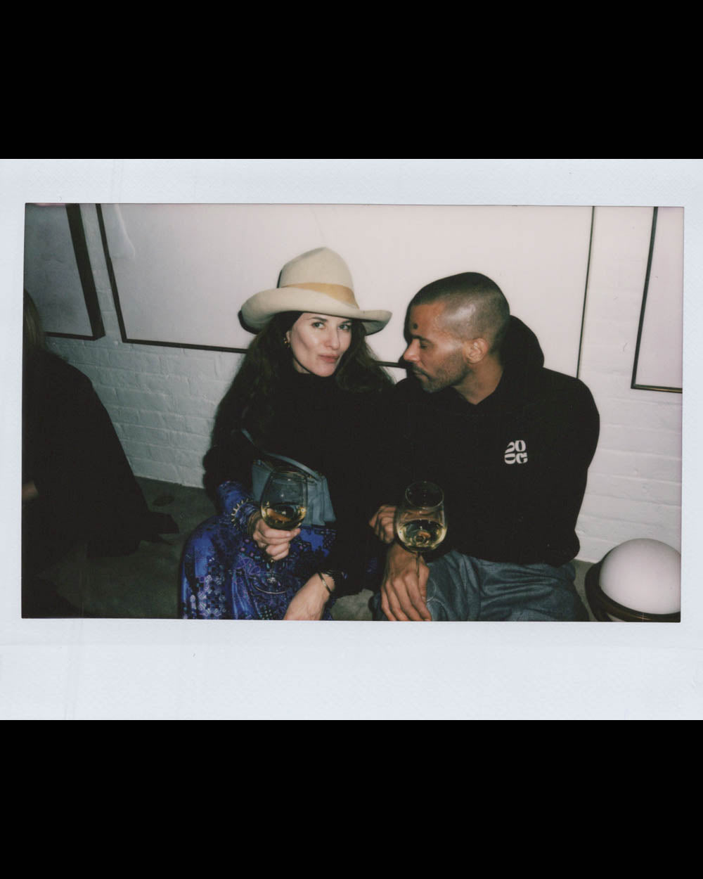A woman wearing a hat and a man in dark clothing sit close, each holding a glass of white wine indoors.