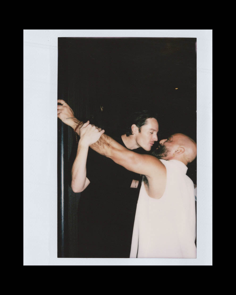 Two men facing each other closely, one with a beard and sleeveless top, holding the other's wrist in a dark setting.
