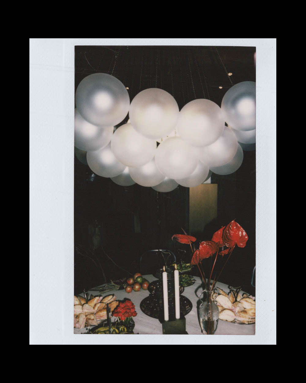 Cluster of glowing white balloons suspended over a table set with candles, flowers, and various foods in dim lighting.