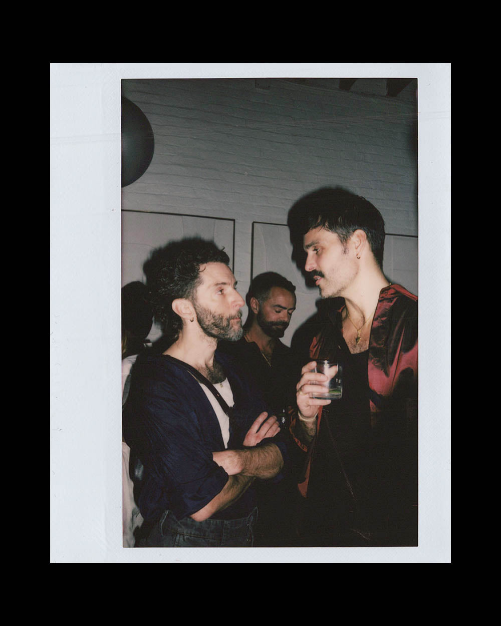 Two men standing close indoors, one holding a drink, engaged in a quiet conversation with a third man in the background.