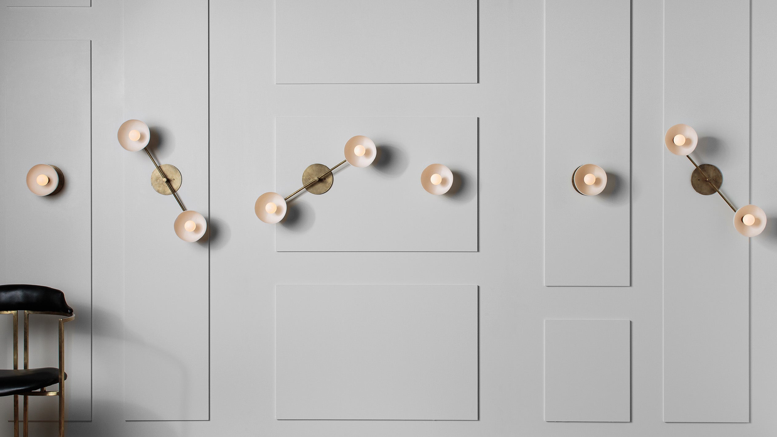 Wall-mounted modern brass sconces with circular frosted glass shades on a white paneled wall above a black and brass stool